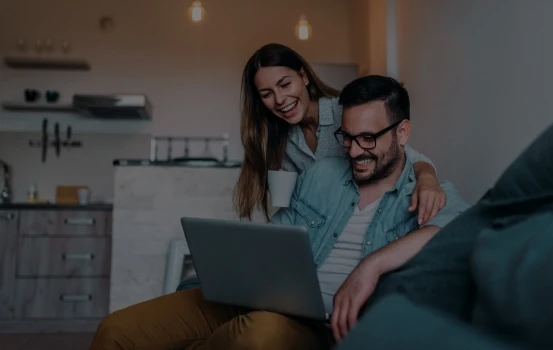 Imagem de um casal feliz sentados no sofá olhando para a tela de um notebook